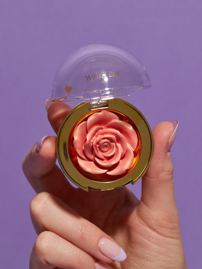 Rosé -- WinkyLux-Digital-Website-Images-Carousel-CheekyRoseHighlighter-1 hand holding up cheeky rose cream highlighter on purple background