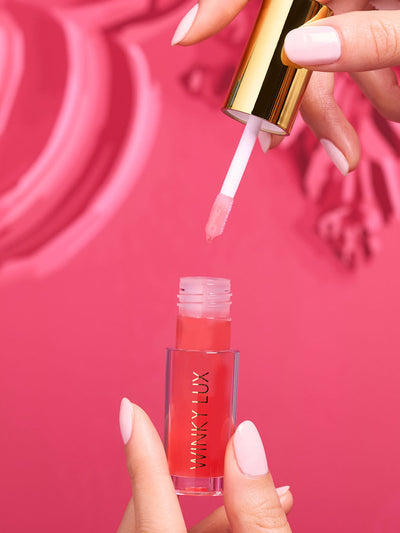 peach whisper -- model hand pulling applicator out of lip oil component on pink background